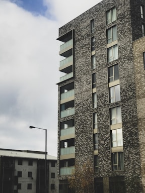 A modern, multi-story apartment building with a textured brick facade and large glass windows. Balconies with glass railings are visible on the side of the structure. The sky is overcast with patches of blue peeking through the clouds, and there's a streetlight in front of the building.