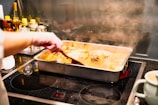 A person stirs a baking dish filled with a steaming, liquid-based dish on a stovetop. Several bottles and containers, likely cooking ingredients, are arranged in the background.