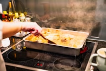A person stirs a baking dish filled with a steaming, liquid-based dish on a stovetop. Several bottles and containers, likely cooking ingredients, are arranged in the background.