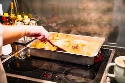 A person stirs a baking dish filled with a steaming, liquid-based dish on a stovetop. Several bottles and containers, likely cooking ingredients, are arranged in the background.