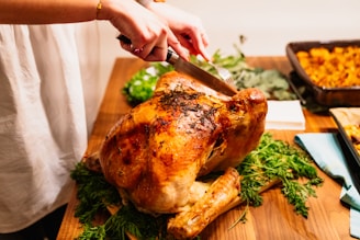 A roasted turkey being carved by a person using a knife and fork on a wooden table. The turkey is garnished with herbs, and there are trays of side dishes visible in the background. A hand is seen holding the carving tools.