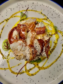 A gourmet dish featuring lobster pieces arranged on a beautifully patterned plate. The lobster is complemented by sprinkles of poppy seeds and is garnished with vibrant sauces and microgreens.