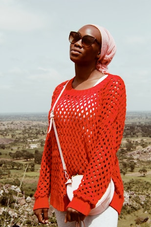 A person wearing a red, knitted sweater, sunglasses, and a pink headscarf stands outdoors against a backdrop of an expansive natural landscape with green fields and scattered trees. The person is also carrying a small, white crossbody bag.