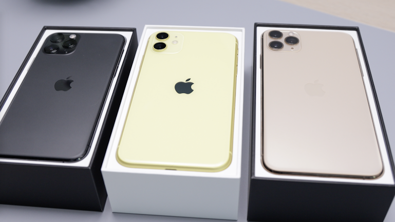 Three smartphones are placed in their respective boxes. The phones are different colors: black, gold, and yellow. Each phone features a prominent Apple logo on the back, with the black and gold devices having three camera lenses, while the yellow phone has two.
