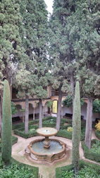 A tranquil garden with a central stone fountain surrounded by manicured hedges and tall cypress trees. The courtyard features arched walkways leading to different sections, with benches placed along the walls. The greenery is lush and well-maintained, contributing to a serene and peaceful atmosphere.