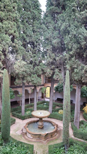 A tranquil garden with a central stone fountain surrounded by manicured hedges and tall cypress trees. The courtyard features arched walkways leading to different sections, with benches placed along the walls. The greenery is lush and well-maintained, contributing to a serene and peaceful atmosphere.