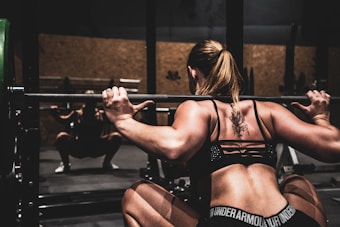 A person with a tattoo on their back is performing a squat with a barbell in a gym environment. The focus is on their muscular back and arms as they hold the barbell. The lighting is dim, creating shadows and highlighting the intensity of the workout.