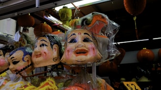 A display of large colorful masks, traditionally crafted with exaggerated facial features, is seen hanging in a market setting. These masks are encased in plastic and are surrounded by red decorative lanterns and various other items, suggesting a festive or cultural context. The scene is brightly lit, highlighting the intricate details and vibrant colors of the masks.