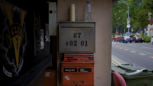 A metallic mailbox with an address labeled '67 #02 01' mounted on a vertical surface. On top of the mailbox, there are two containers, one resembling a water bottle. Below the mailbox, a red postbox labeled '#03-01' is affixed. The background features a street scene with cars, greenery, and sidewalks. There is signage with the text 'Club 3' on a black backdrop to the left, adding to the urban environment.