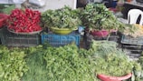 An assortment of fresh vegetables and greens are displayed in baskets and crates. The focal point includes vibrant red radishes and a variety of leafy greens, all set up on a makeshift stall made of stacked crates. A white plastic chair is visible in the background.