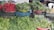 An assortment of fresh vegetables and greens are displayed in baskets and crates. The focal point includes vibrant red radishes and a variety of leafy greens, all set up on a makeshift stall made of stacked crates. A white plastic chair is visible in the background.
