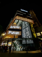 A modern, illuminated building at night with prominent signage that reads 'CapitaLand' and 'Funan'. The exterior features a vertical garden and intricate lighting patterns. People are gathered at the entrance, and there are visible escalators leading inside.
