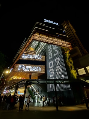 A modern, illuminated building at night with prominent signage that reads 'CapitaLand' and 'Funan'. The exterior features a vertical garden and intricate lighting patterns. People are gathered at the entrance, and there are visible escalators leading inside.