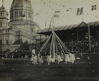 A group of children in white dresses are dancing around a maypole with long ribbons in a large outdoor area. Behind them, a grand building with a domed roof and decorative towers stands, resembling an exhibition hall. The scene is bustling with a large crowd of spectators seated in a covered stand. Flags hang overhead, creating a festive atmosphere.