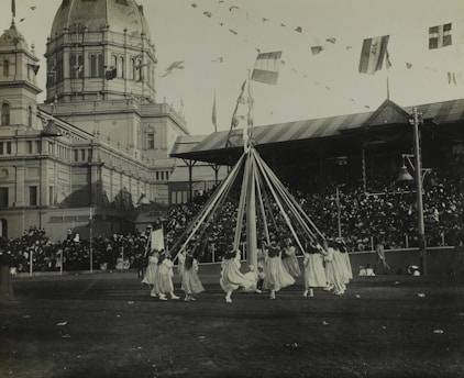 A group of children in white dresses are dancing around a maypole with long ribbons in a large outdoor area. Behind them, a grand building with a domed roof and decorative towers stands, resembling an exhibition hall. The scene is bustling with a large crowd of spectators seated in a covered stand. Flags hang overhead, creating a festive atmosphere.