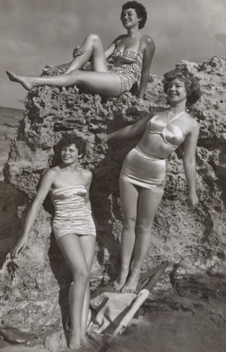 grayscale photo of three women standing and sitting on rocks