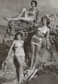 grayscale photo of three women standing and sitting on rocks