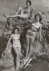 grayscale photo of three women standing and sitting on rocks