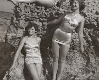 grayscale photo of three women standing and sitting on rocks