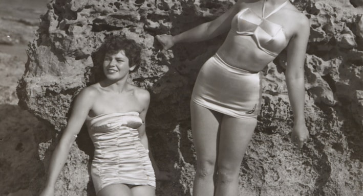 grayscale photo of three women standing and sitting on rocks