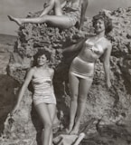 grayscale photo of three women standing and sitting on rocks