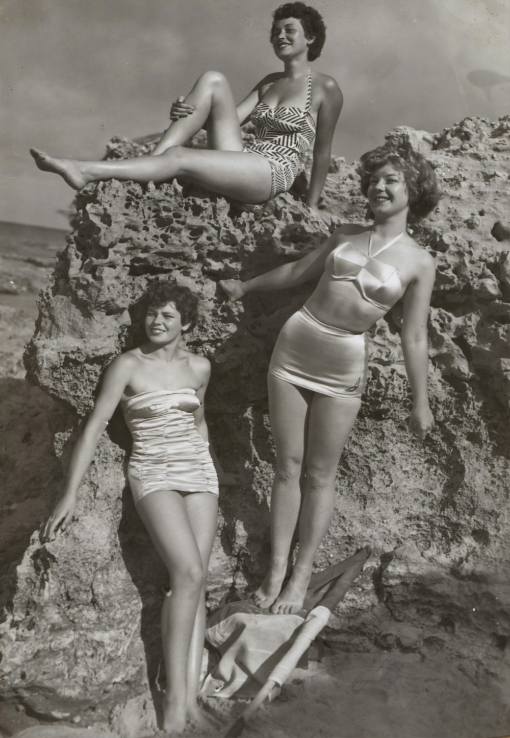 grayscale photo of three women standing and sitting on rocks