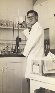 A person wearing a lab coat is standing in a laboratory. They are adjusting a piece of equipment with a tall glass tube. Various glass bottles and beakers are arranged on the counter and shelves behind them. A box labeled 'Photostat' and a cloth are on a nearby table.
