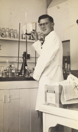 A person wearing a lab coat is standing in a laboratory. They are adjusting a piece of equipment with a tall glass tube. Various glass bottles and beakers are arranged on the counter and shelves behind them. A box labeled 'Photostat' and a cloth are on a nearby table.
