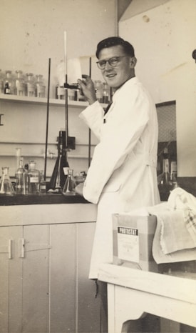 A person wearing a lab coat is standing in a laboratory. They are adjusting a piece of equipment with a tall glass tube. Various glass bottles and beakers are arranged on the counter and shelves behind them. A box labeled 'Photostat' and a cloth are on a nearby table.