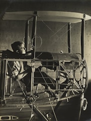 A person sits in an early aviation machine, surrounded by a complex framework and wires. The structure has a skeletal frame, reminiscent of vintage airplane designs. The lighting is dramatic, casting shadows that highlight the intricate details of the machine and the thoughtful expression of the person.