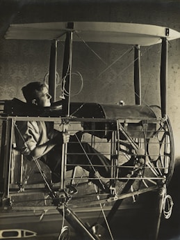 A person sits in an early aviation machine, surrounded by a complex framework and wires. The structure has a skeletal frame, reminiscent of vintage airplane designs. The lighting is dramatic, casting shadows that highlight the intricate details of the machine and the thoughtful expression of the person.