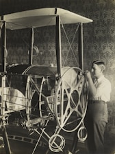 A young person is examining or working on an early model of an aircraft or contraption with large wheels and a framework structure. The setting appears to be indoors, with a detailed, patterned wallpaper in the background.
