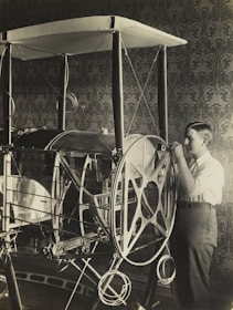 A young person is examining or working on an early model of an aircraft or contraption with large wheels and a framework structure. The setting appears to be indoors, with a detailed, patterned wallpaper in the background.