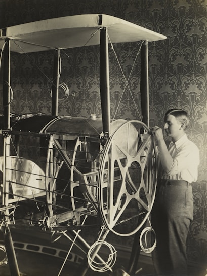A young person is examining or working on an early model of an aircraft or contraption with large wheels and a framework structure. The setting appears to be indoors, with a detailed, patterned wallpaper in the background.