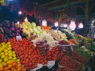 A vibrant market scene featuring an abundant display of fresh fruits and vegetables. Large piles of tomatoes, oranges, cabbages, and eggplants are meticulously arranged. A variety of peppers hang overhead, adding color and texture. The area is well-lit with several bright light bulbs hanging above, creating a lively and welcoming atmosphere. A person is visible on the right, partially obscured by the produce.
