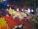 A vibrant market scene featuring an abundant display of fresh fruits and vegetables. Large piles of tomatoes, oranges, cabbages, and eggplants are meticulously arranged. A variety of peppers hang overhead, adding color and texture. The area is well-lit with several bright light bulbs hanging above, creating a lively and welcoming atmosphere. A person is visible on the right, partially obscured by the produce.