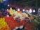 A vibrant market scene featuring an abundant display of fresh fruits and vegetables. Large piles of tomatoes, oranges, cabbages, and eggplants are meticulously arranged. A variety of peppers hang overhead, adding color and texture. The area is well-lit with several bright light bulbs hanging above, creating a lively and welcoming atmosphere. A person is visible on the right, partially obscured by the produce.