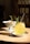 A glass of yellow drink with a straw garnished with a rosemary sprig and a slice of lemon is placed on a saucer on a wooden table. Next to it, there's a white pedestal bowl containing a small portion of food. The background features a chair and a dimly lit room.
