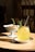A glass of yellow drink with a straw garnished with a rosemary sprig and a slice of lemon is placed on a saucer on a wooden table. Next to it, there's a white pedestal bowl containing a small portion of food. The background features a chair and a dimly lit room.