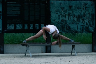 A person wearing a white shirt, black shorts, and sneakers is stretching on a bench. Their body is bent over with arms extended, and they appear to be listening to music with earphones. The background features panels with historical images and text, and there is grass visible behind the panels.