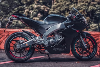 A sleek black motorcycle is parked on a road surface, featuring a sporty design with aerodynamic contours. The background consists of blurred green foliage and an orange barrier for contrast. The motorcycle has a prominent 'aprilia' logo on the side, and its metallic components and precise engineering are visible.