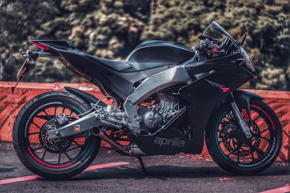 A sleek black motorcycle is parked on a road surface, featuring a sporty design with aerodynamic contours. The background consists of blurred green foliage and an orange barrier for contrast. The motorcycle has a prominent 'aprilia' logo on the side, and its metallic components and precise engineering are visible.