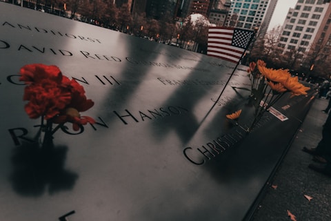 Engraved names on a memorial plaque are surrounded by red and yellow flowers, with an American flag placed among them. The scene is set against a backdrop of autumn trees and city buildings, evoking a contemplative atmosphere.