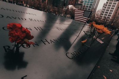 Engraved names on a memorial plaque are surrounded by red and yellow flowers, with an American flag placed among them. The scene is set against a backdrop of autumn trees and city buildings, evoking a contemplative atmosphere.
