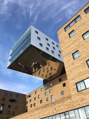 A modern architectural structure featuring a large, rectangular building extension that appears to be partially suspended in the air. The extension is covered in reflective glass and metal, creating an illusion of floating above the main brick building. Various windows of different sizes are present on both the extension and the main building. Clear blue sky forms the backdrop.