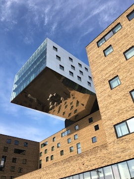 A modern architectural structure featuring a large, rectangular building extension that appears to be partially suspended in the air. The extension is covered in reflective glass and metal, creating an illusion of floating above the main brick building. Various windows of different sizes are present on both the extension and the main building. Clear blue sky forms the backdrop.