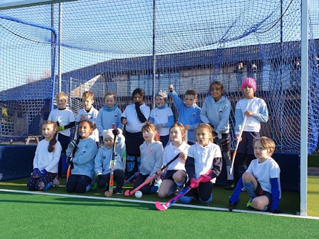 A group of young children are gathered on a sports field, posing in front of a blue net. They are dressed in sports attire, holding field hockey sticks, and appear to be part of a team. The sun is shining brightly, casting shadows on the field.