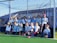 A group of young children are gathered on a sports field, posing in front of a blue net. They are dressed in sports attire, holding field hockey sticks, and appear to be part of a team. The sun is shining brightly, casting shadows on the field.