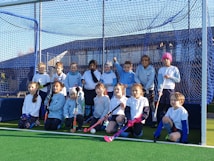 A group of young children are gathered on a sports field, posing in front of a blue net. They are dressed in sports attire, holding field hockey sticks, and appear to be part of a team. The sun is shining brightly, casting shadows on the field.
