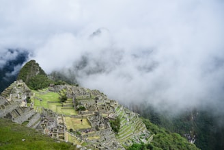 An aerial view of ancient stone structures nestled in the mountains, partially covered by mist and surrounded by lush green vegetation. The stone terraces and ruins indicate an archaeological site, with pathways and platforms visible throughout the landscape.